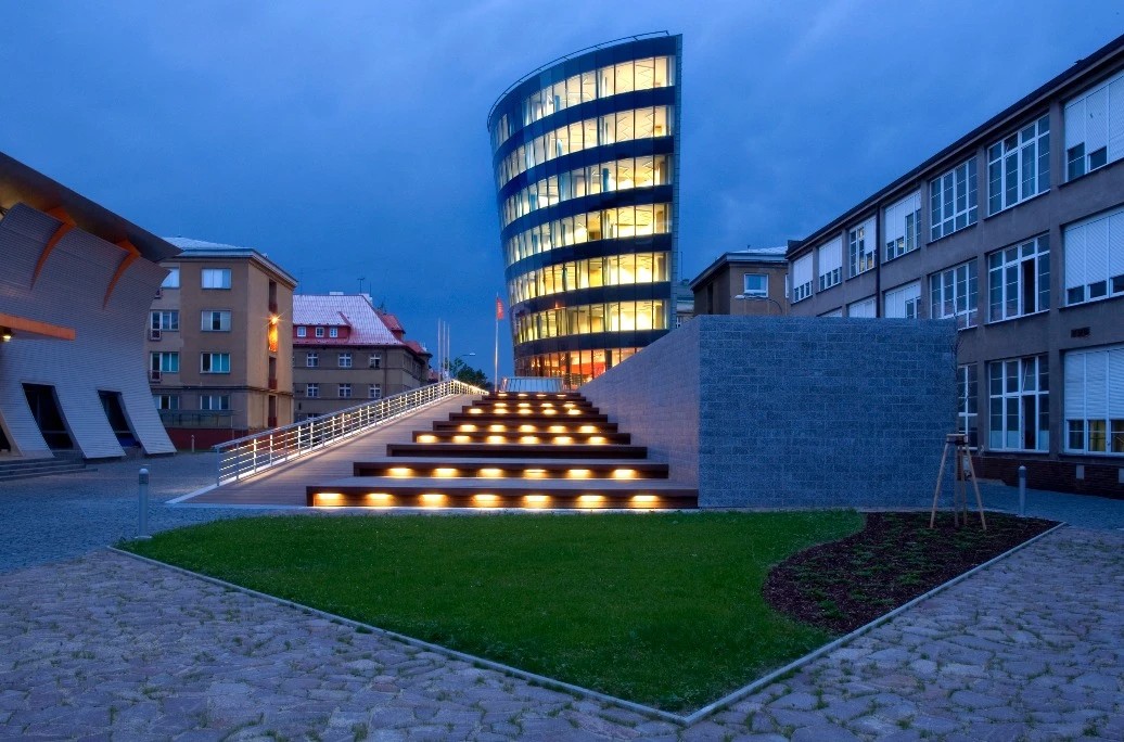 A curved glass building at Technical University of Liberec lit from within at dusk, illuminated steps rising from a cobbled plaza